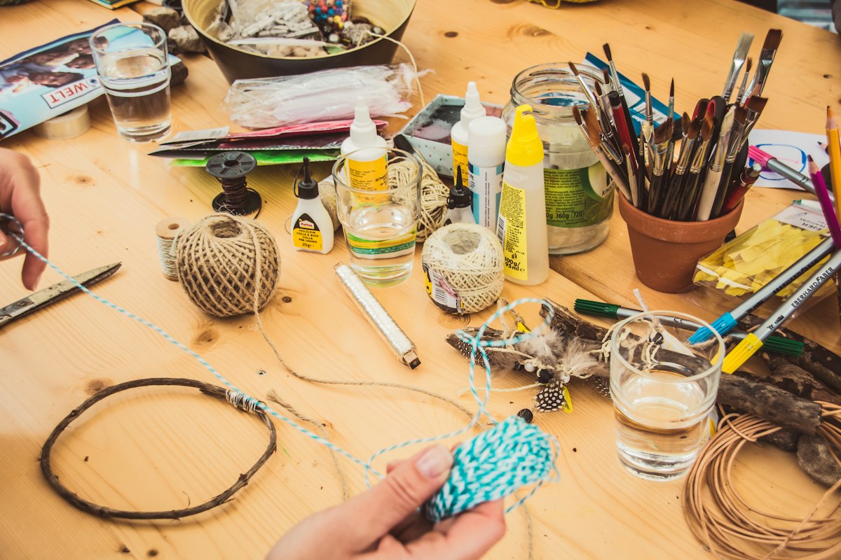 Mesa de trabajo de manualidades con ovillos de hilo, botellas de pegamento, pinceles en una maceta de terracota, lápices de colores y unas manos sosteniendo cordel azul y blanco.